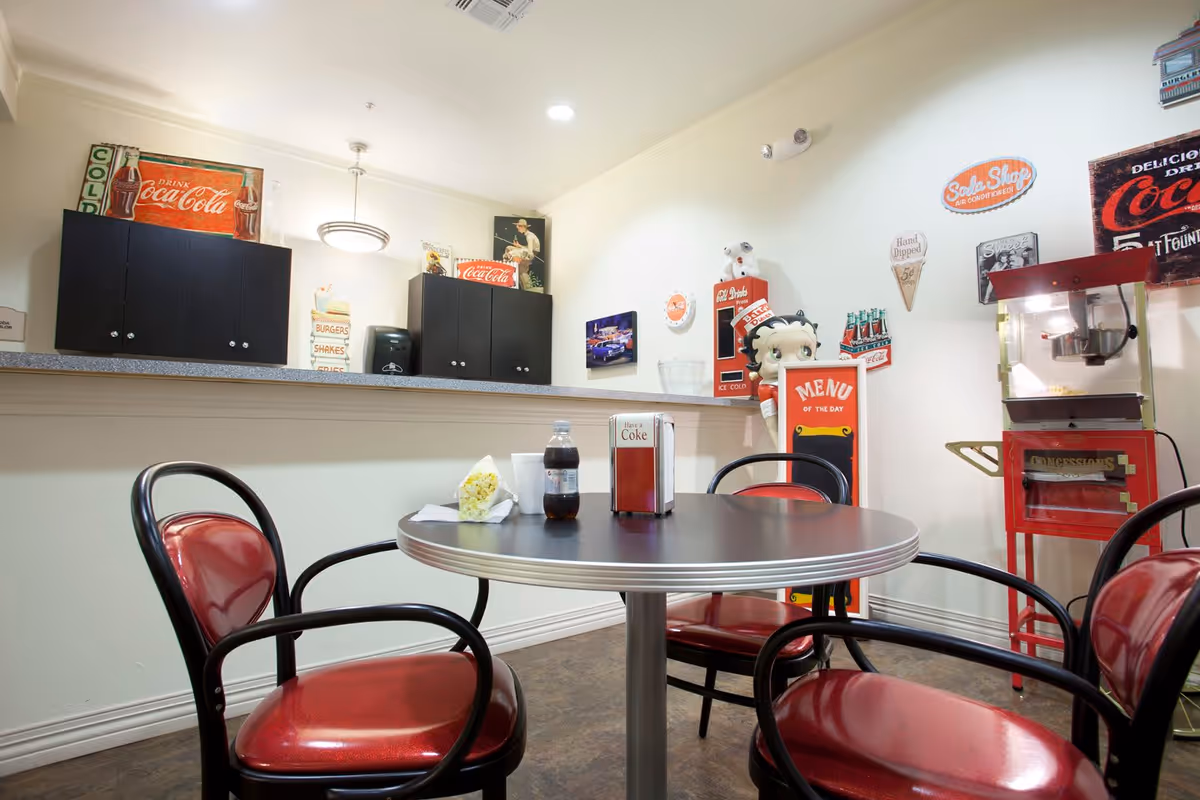 A retro-style dining area with a round table surrounded by four red and black chairs. On the table, there is a bottle of soda, a napkin dispenser, a cup, and a small bag of popcorn. The background features a counter with black cabinets and vintage Coca-Cola signs. There is also a popcorn machine and a Betty Boop figure holding a 'Menu of the Day' sign, along with various nostalgic wall decorations.