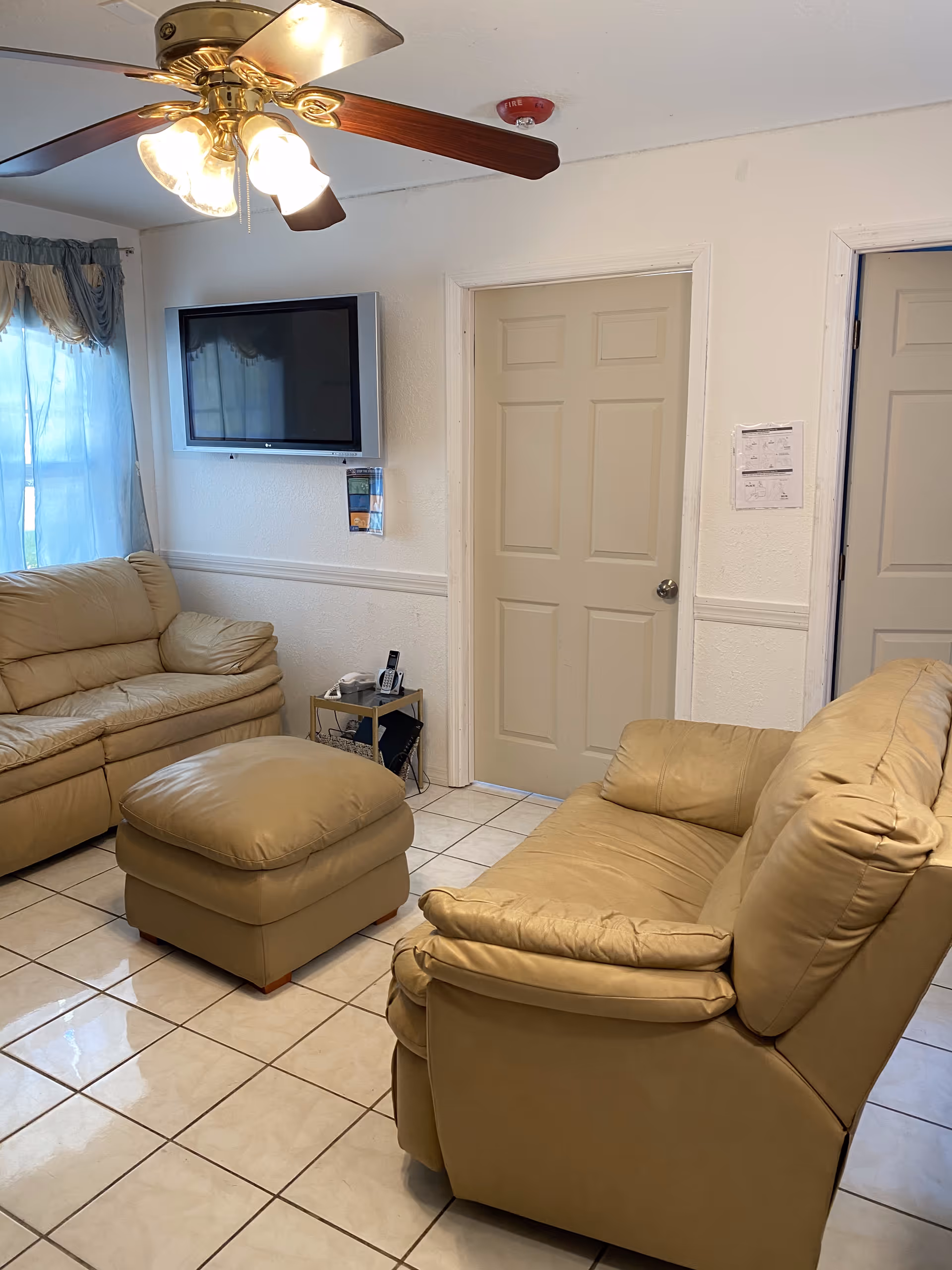 A cozy living room with beige leather sofas and a matching ottoman on a tiled floor. A ceiling fan with lights is mounted on the ceiling. A flat-screen TV is mounted on the wall above a small side table holding a telephone and other items. Two closed doors are visible on the wall opposite the sofas, and a window with blue curtains lets in natural light.