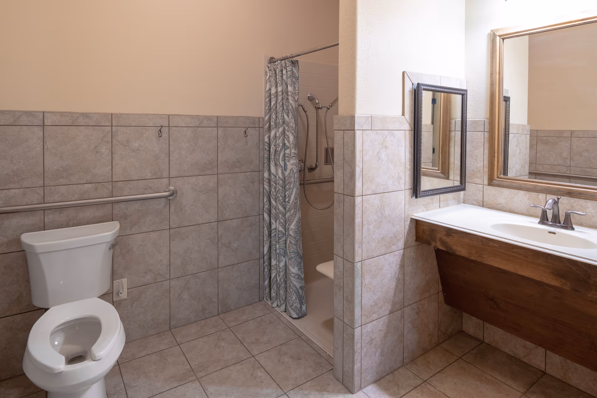 A senior living facility bathroom featuring a white toilet with safety grab bars, a tiled floor and walls, a walk-in shower with a patterned shower curtain, and a sink with a large mirror above it.