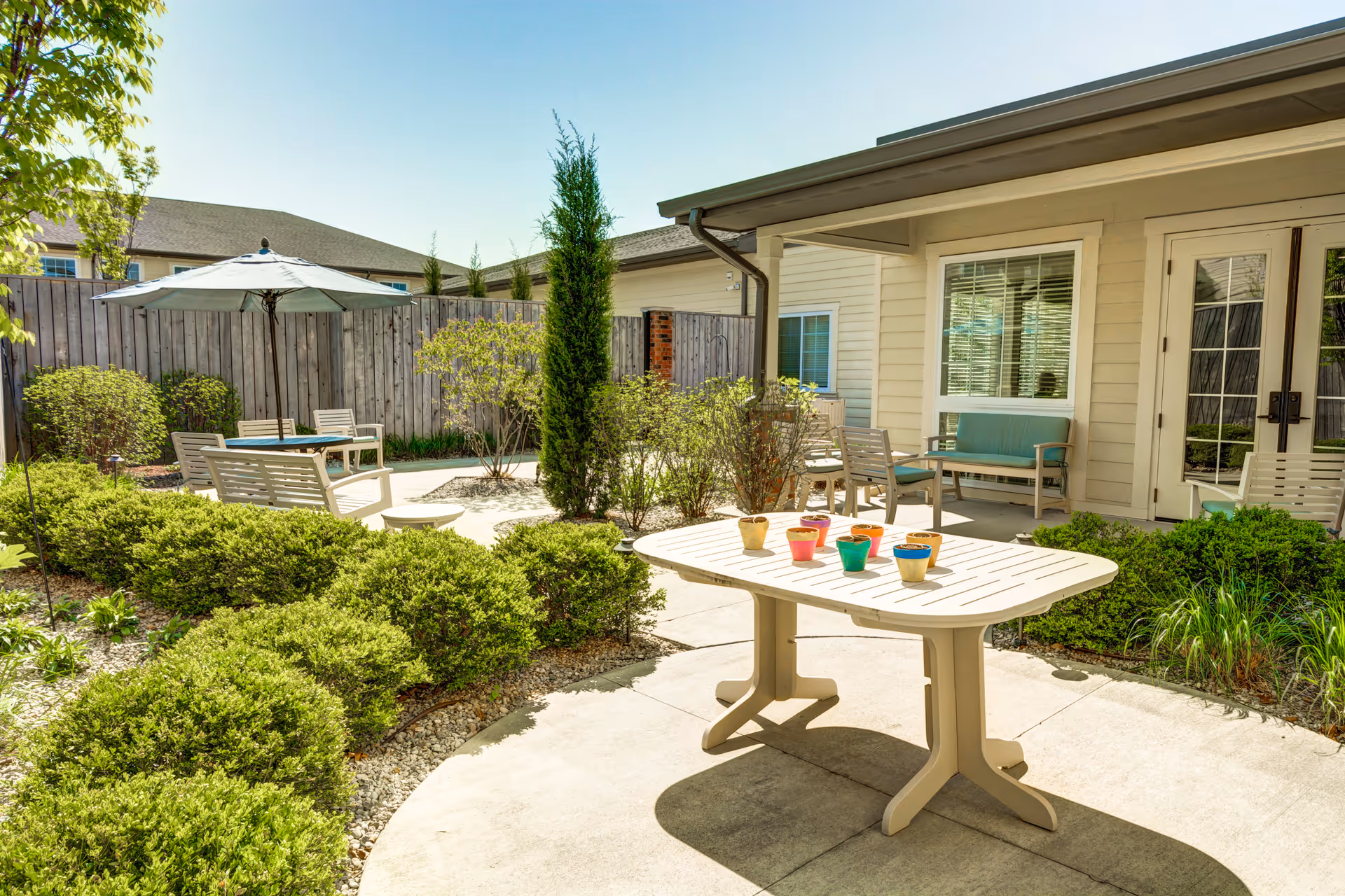 Outdoor patio area at Traditions of West Chester featuring a white table with colorful small pots, surrounded by green bushes and plants. There are several chairs and benches with cushions, a large umbrella providing shade, and a wooden fence enclosing the space. The building exterior is visible with windows and a door leading to the patio.