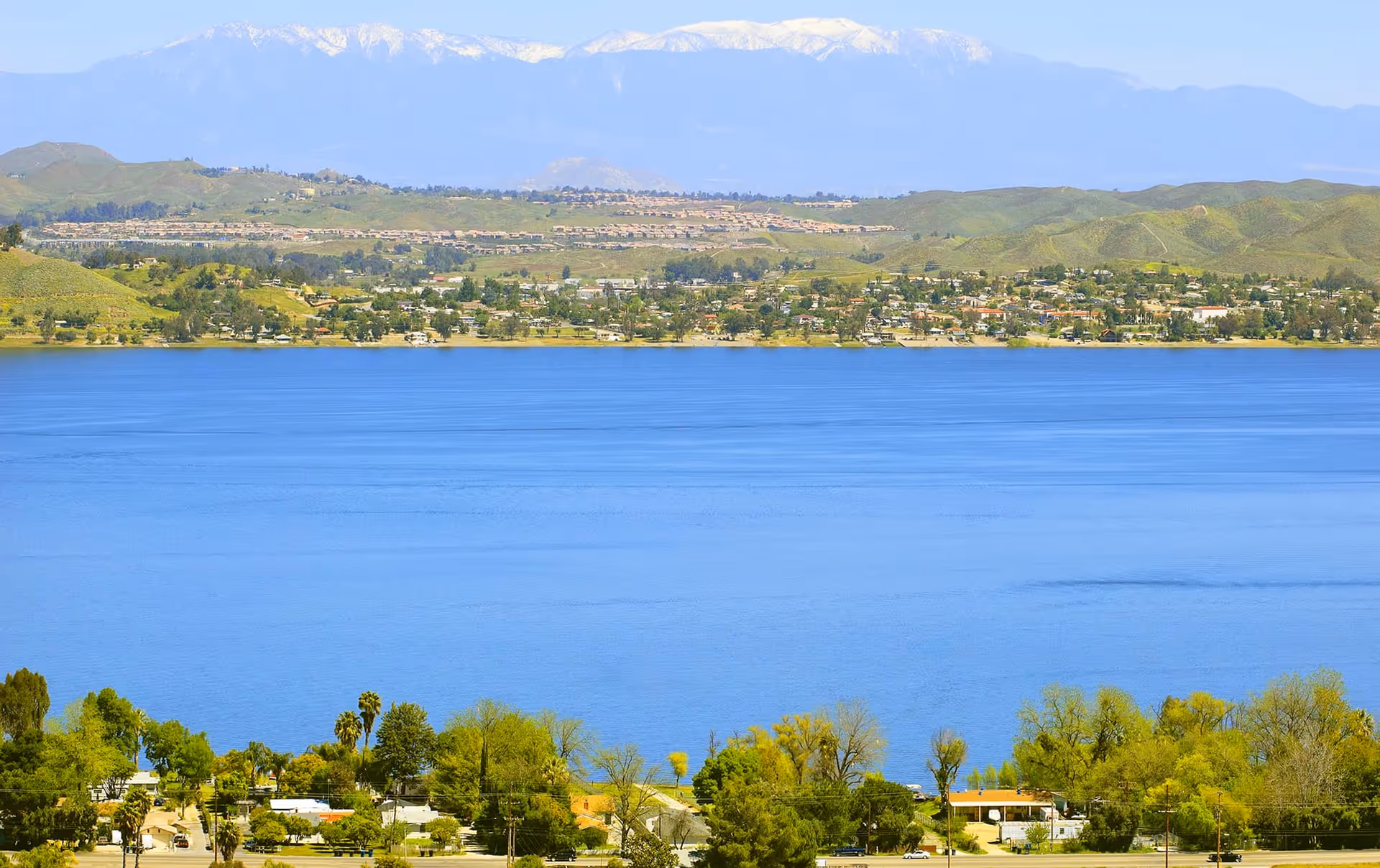 Wide view of a blue lake with a shoreline of houses and green hills and snow-capped mountains in the distance.