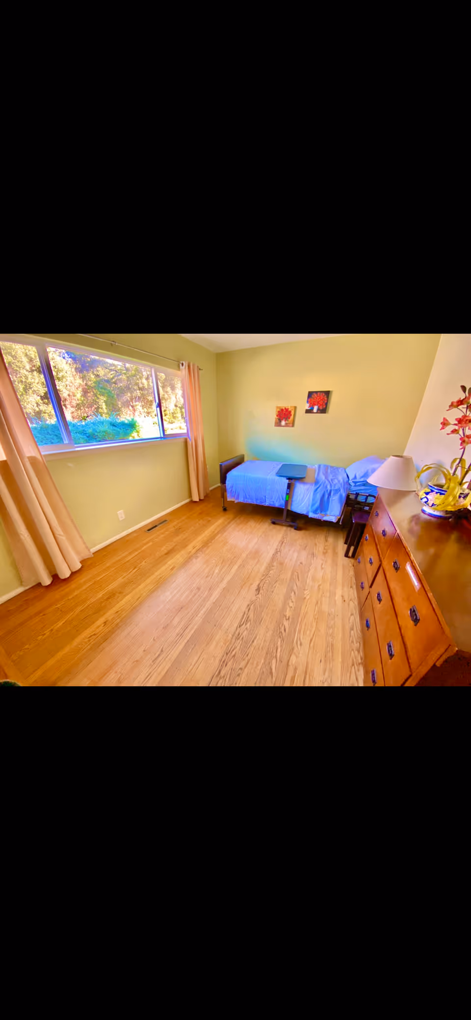 A simple bedroom with light green walls and wooden floor. There is a single bed with blue bedding against the far wall, a small overbed table, a wooden dresser with a lamp and decorative items, and two framed floral pictures on the wall. A large window with beige curtains lets in natural light and shows greenery outside.