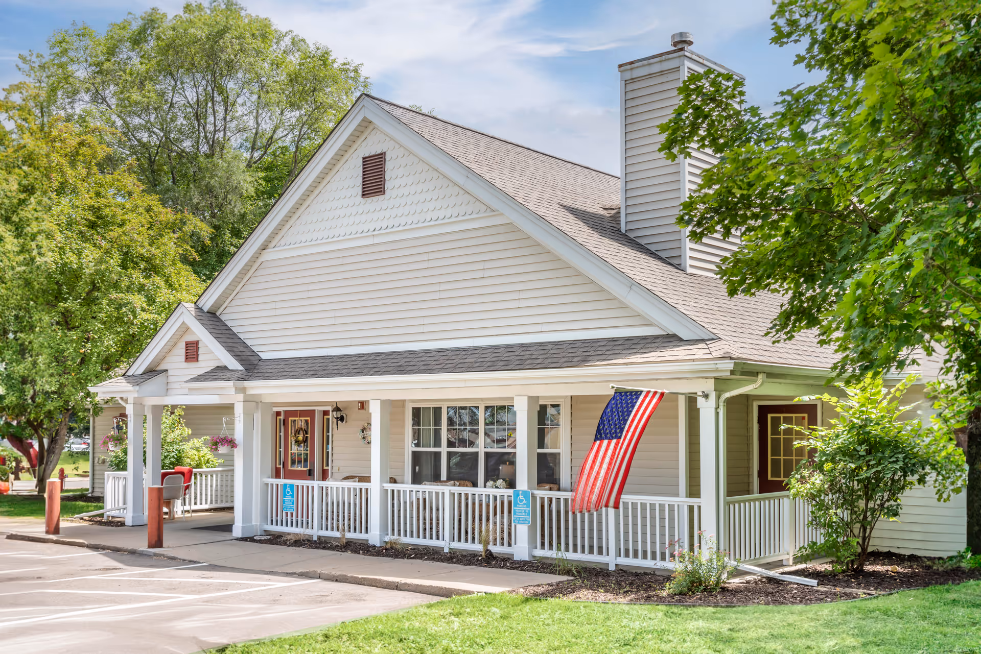 Exterior view of a single-story building with white siding and a gray shingled roof. The building has a covered porch with white railings and an American flag hanging near the entrance. There are trees and greenery surrounding the building, and a parking area is visible in the foreground.