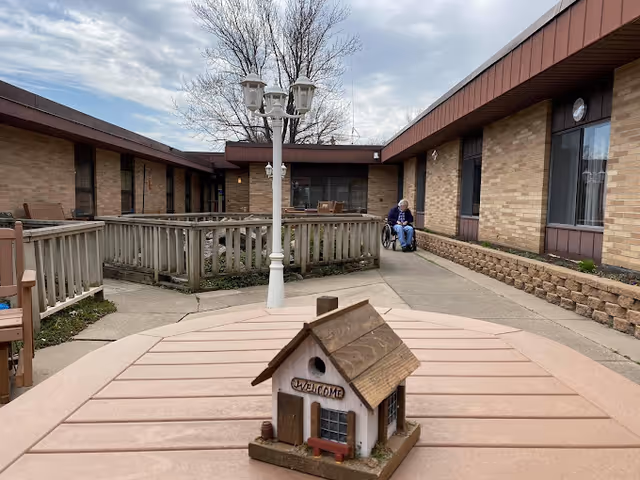 Decorative birdhouse on a table in a senior facility courtyard with a lamp post and a person in a wheelchair by a brick building.