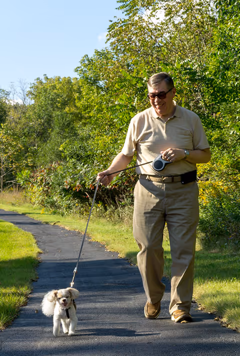 An elderly man wearing sunglasses and casual clothing walks a small white dog on a leash along a paved path surrounded by green trees and grass on a sunny day.