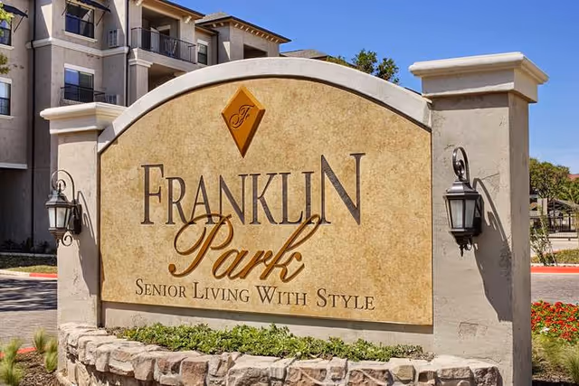 Stone sign at the entrance of Franklin Park Senior Living facility with the text 'Franklin Park Senior Living With Style' and decorative lanterns on either side, with part of the building and clear blue sky in the background.