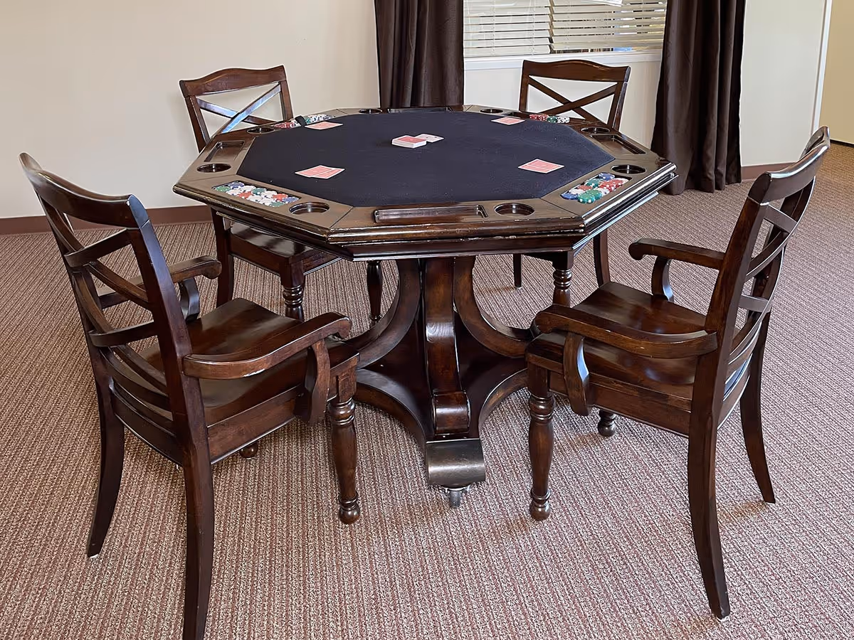 A wooden octagonal poker table with a black felt playing surface, poker chips, and playing cards arranged on it. Four wooden chairs with armrests surround the table. The room has carpeted flooring, a window with blinds, and dark curtains.
