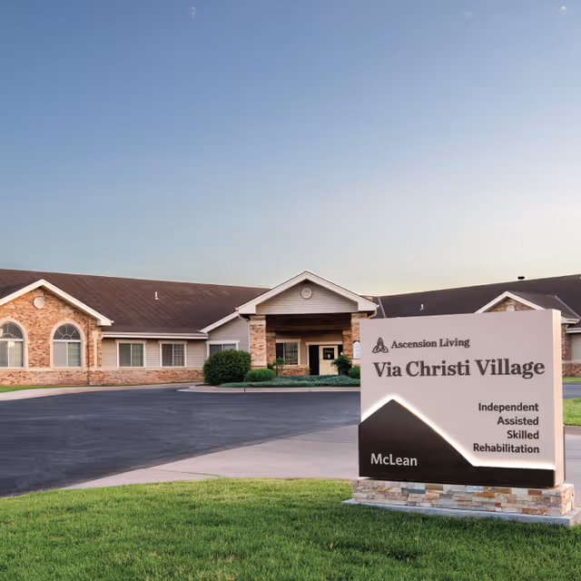 Exterior view of Ascension Living Via Christi Village building with a large sign in the foreground displaying the facility name and services including Independent, Assisted, Skilled, and Rehabilitation. The building has a brick and siding facade with a covered entrance and a paved driveway.