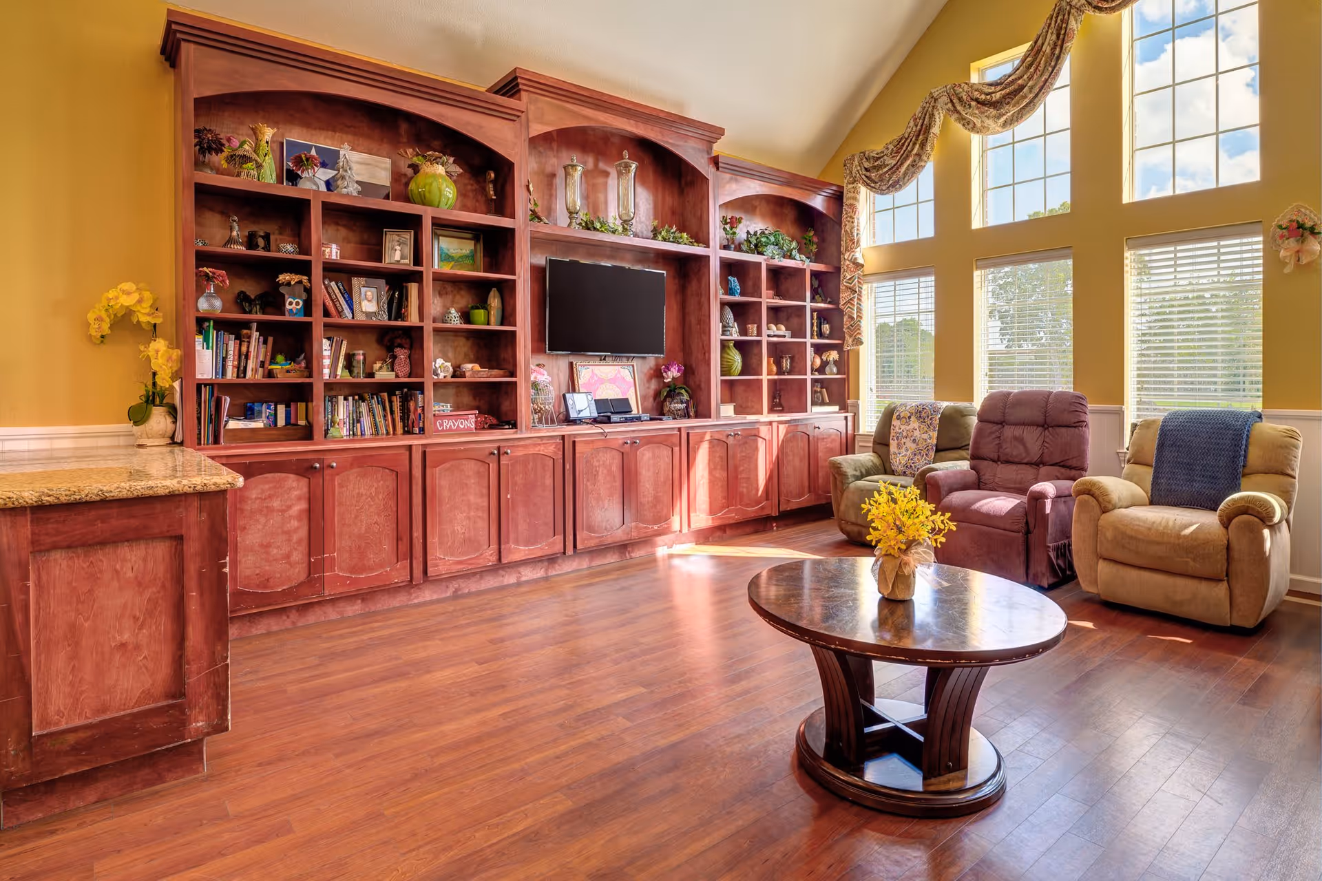 A bright living room with large windows letting in natural light, featuring three cushioned armchairs with blankets draped over them, a wooden coffee table with a small yellow flower arrangement, and a large built-in wooden shelving unit with various decorative items, books, and a mounted flat-screen TV.