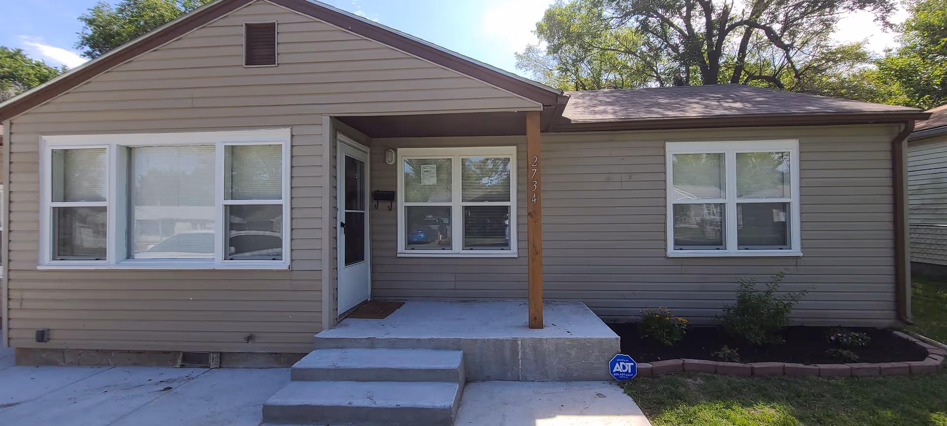 Front exterior view of a single-story house with beige siding, white-framed windows, a small concrete porch with three steps, and a wooden post supporting the porch roof. There is a security sign on the ground near the porch and some small plants along the foundation.