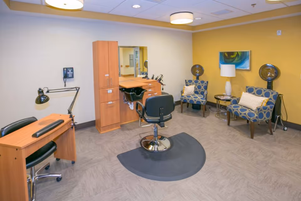 Salon-style room with a central styling chair and sink, a manicure desk, and two patterned chairs with hooded hair dryers against a yellow wall.