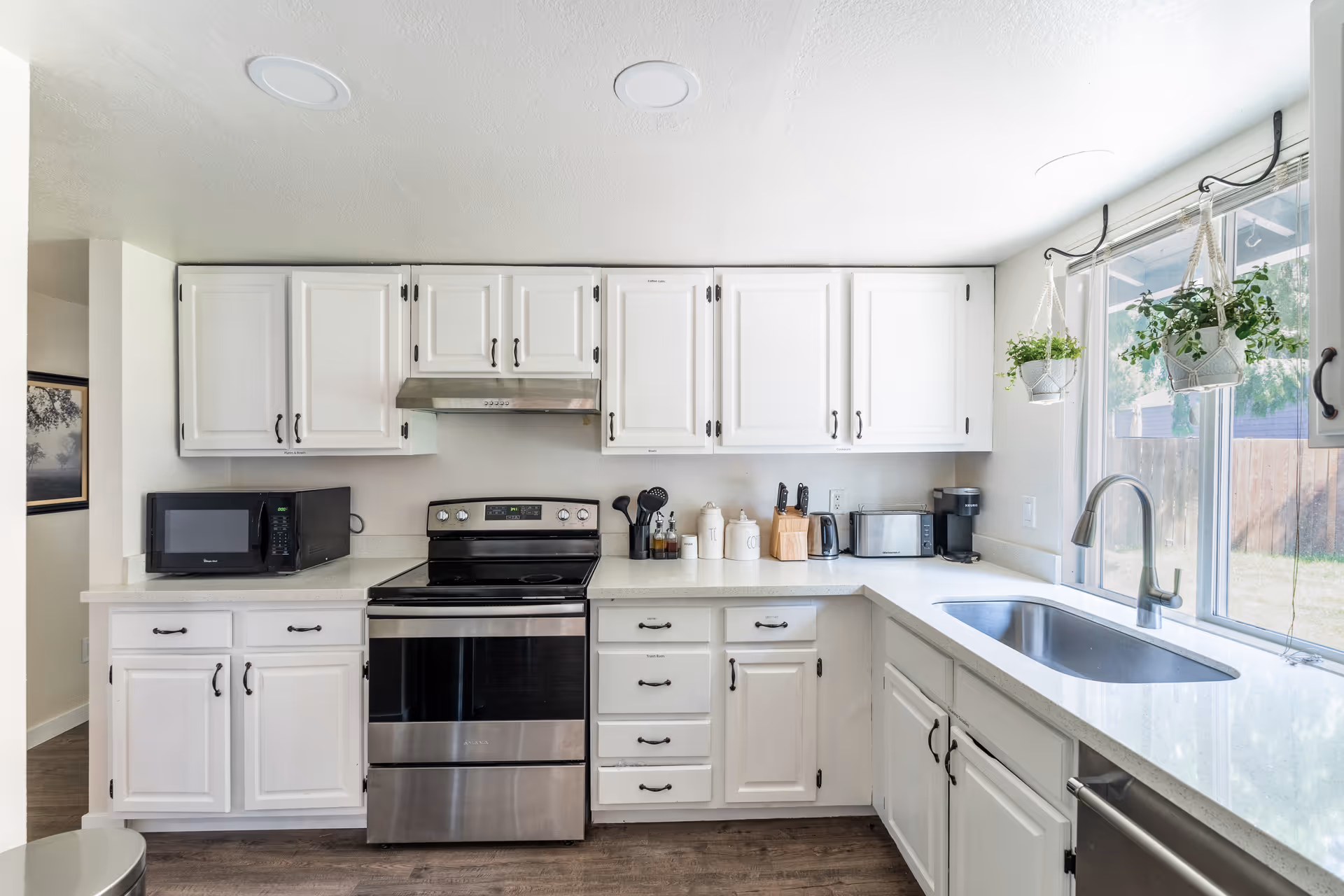 Bright kitchen with white cabinets, stainless steel stove and dishwasher, a microwave, coffee maker, toaster, and hanging plants by a large window overlooking a fenced backyard.