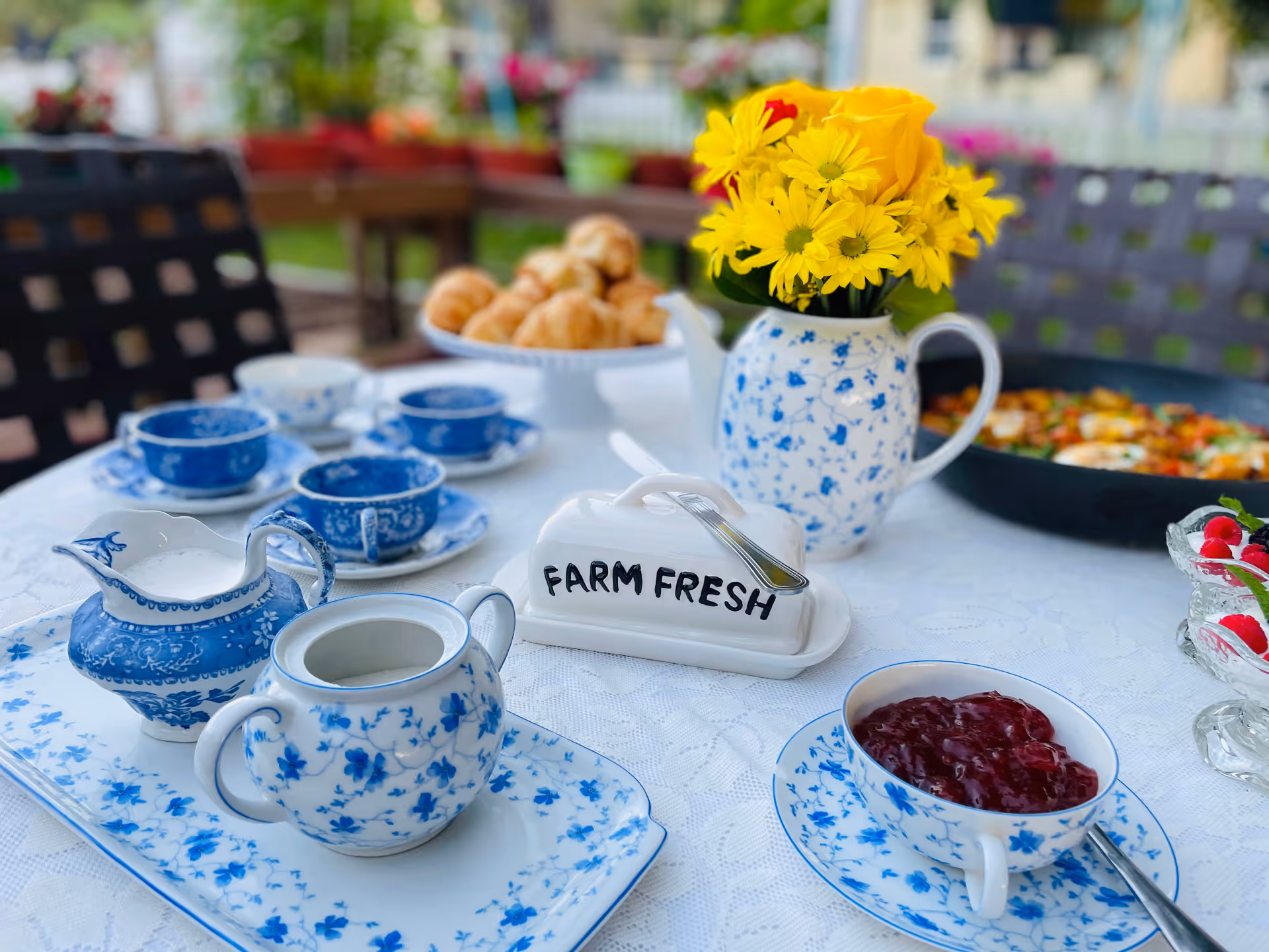 Outdoor table set with blue-and-white china, yellow flowers in a pitcher, croissants, and a 'FARM FRESH' butter dish.