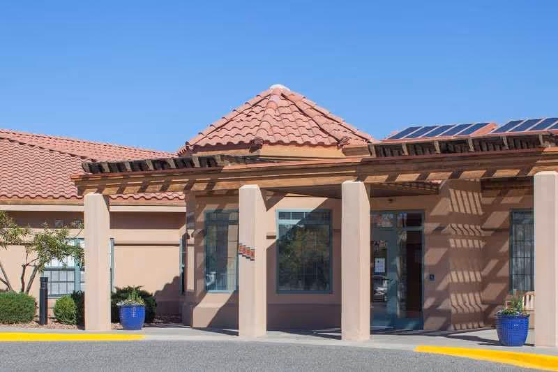 Exterior view of a building with a tiled roof and beige stucco walls, featuring a covered entrance supported by columns and blue potted plants on either side of the entrance.