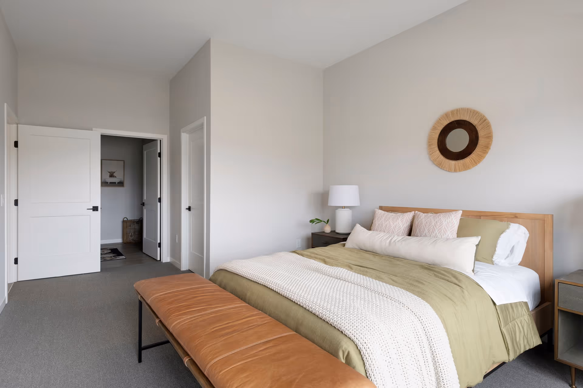 Bright minimalist bedroom with a made bed, a leather bench at the foot, nightstands, and an open doorway.