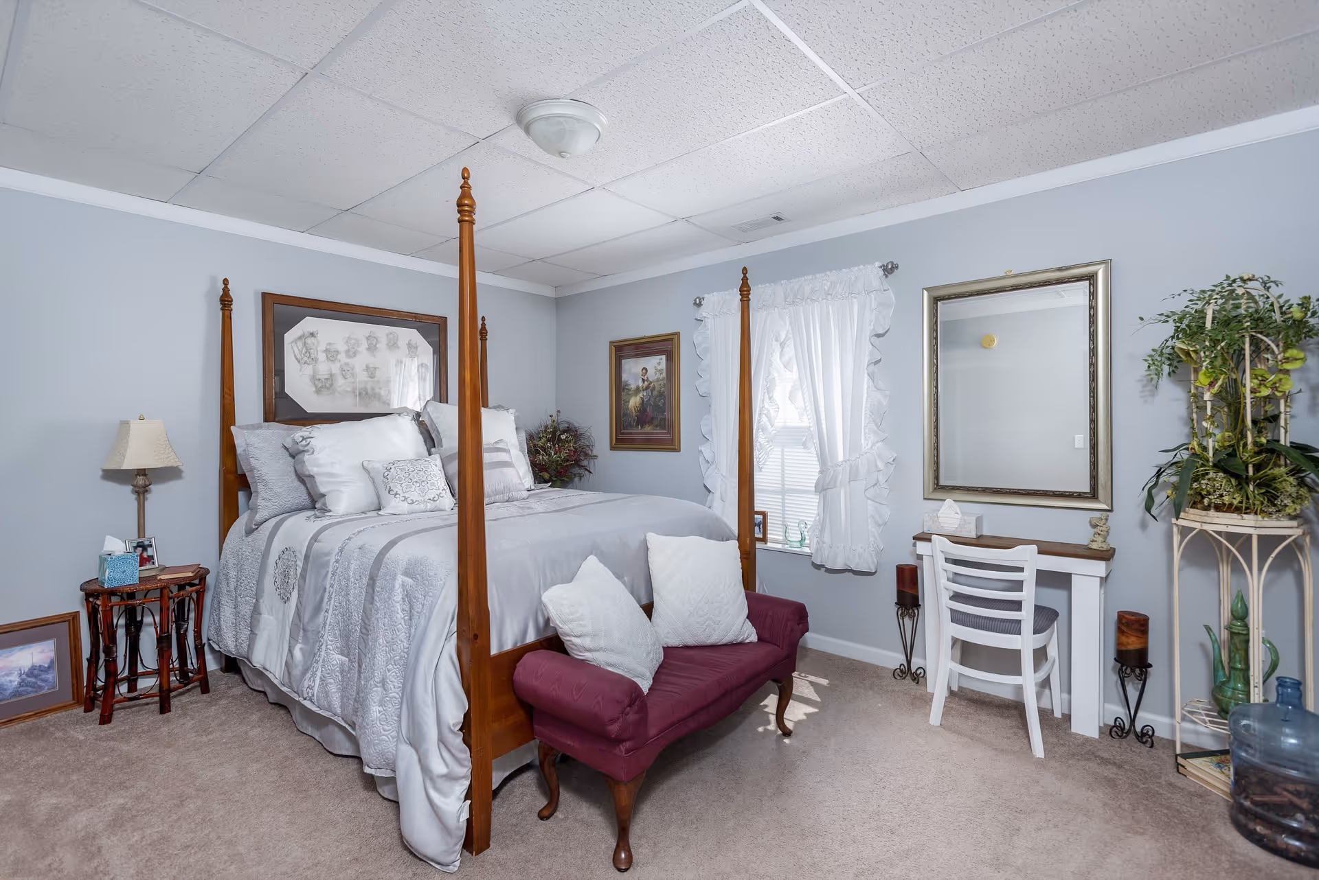 A decorated bedroom with a four-poster bed, layered pillows and bedding, a burgundy bench at the foot, a small desk with mirror, and plants.