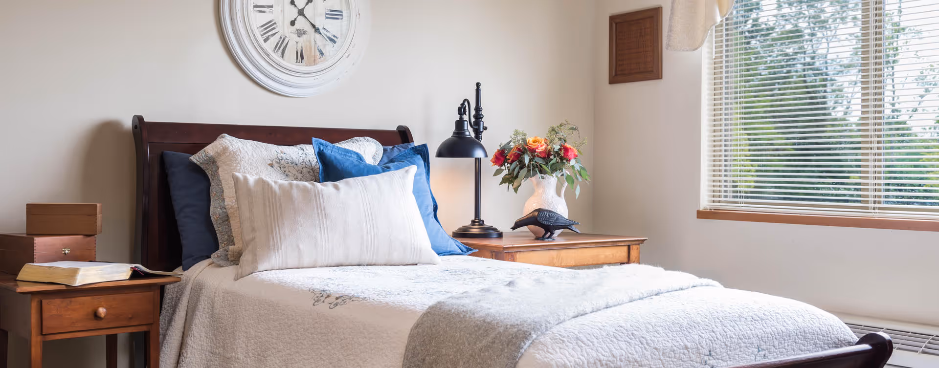 A cozy bedroom with a wooden bed frame, white and blue pillows, and a white quilted bedspread. There is a wooden nightstand on each side of the bed, one with an open book and a small box, and the other with a black lamp and a vase of flowers. A large round clock hangs on the wall above the bed, and a window with blinds lets in natural light.