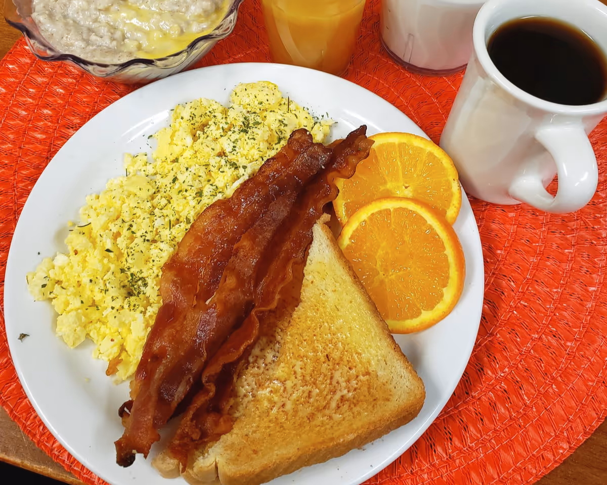 A breakfast plate with scrambled eggs sprinkled with herbs, three strips of crispy bacon, two slices of toasted bread, and two orange slices. Next to the plate are a bowl of oatmeal, a glass of orange juice, a glass of milk, and a white mug filled with black coffee, all placed on an orange woven placemat.