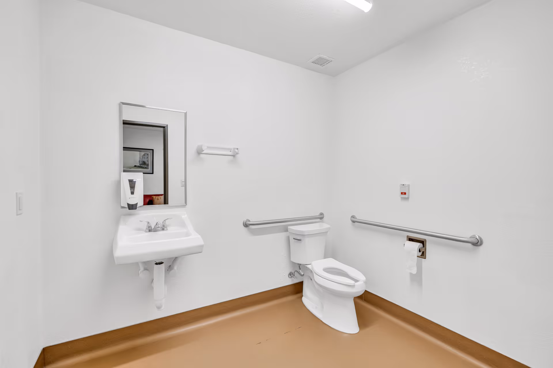 A clean, accessible bathroom with white walls and a brown floor. The bathroom features a white toilet with grab bars on both sides, a white wall-mounted sink with a mirror above it, a soap dispenser attached to the mirror, and a toilet paper holder with a roll of toilet paper. There is also an emergency call button on the wall near the toilet.