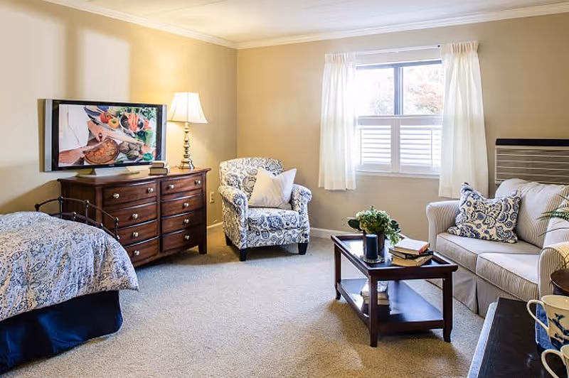 Bright senior living studio with a bed and dresser on the left and a seating area with an armchair, sofa and coffee table by a window.