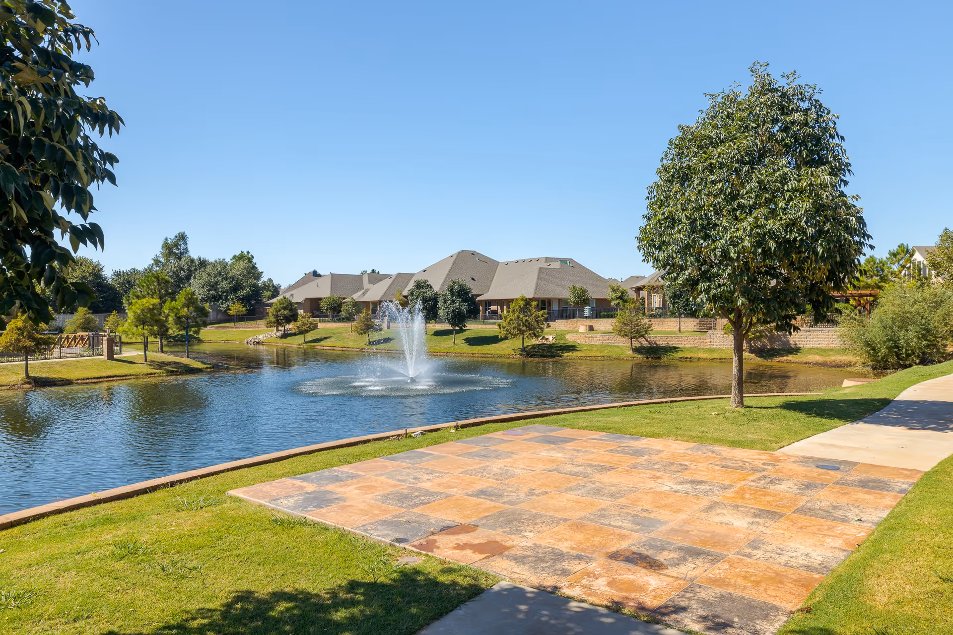 A pond with a central fountain beside a paved patio and walkway, surrounded by grass, trees, and houses under a clear blue sky.