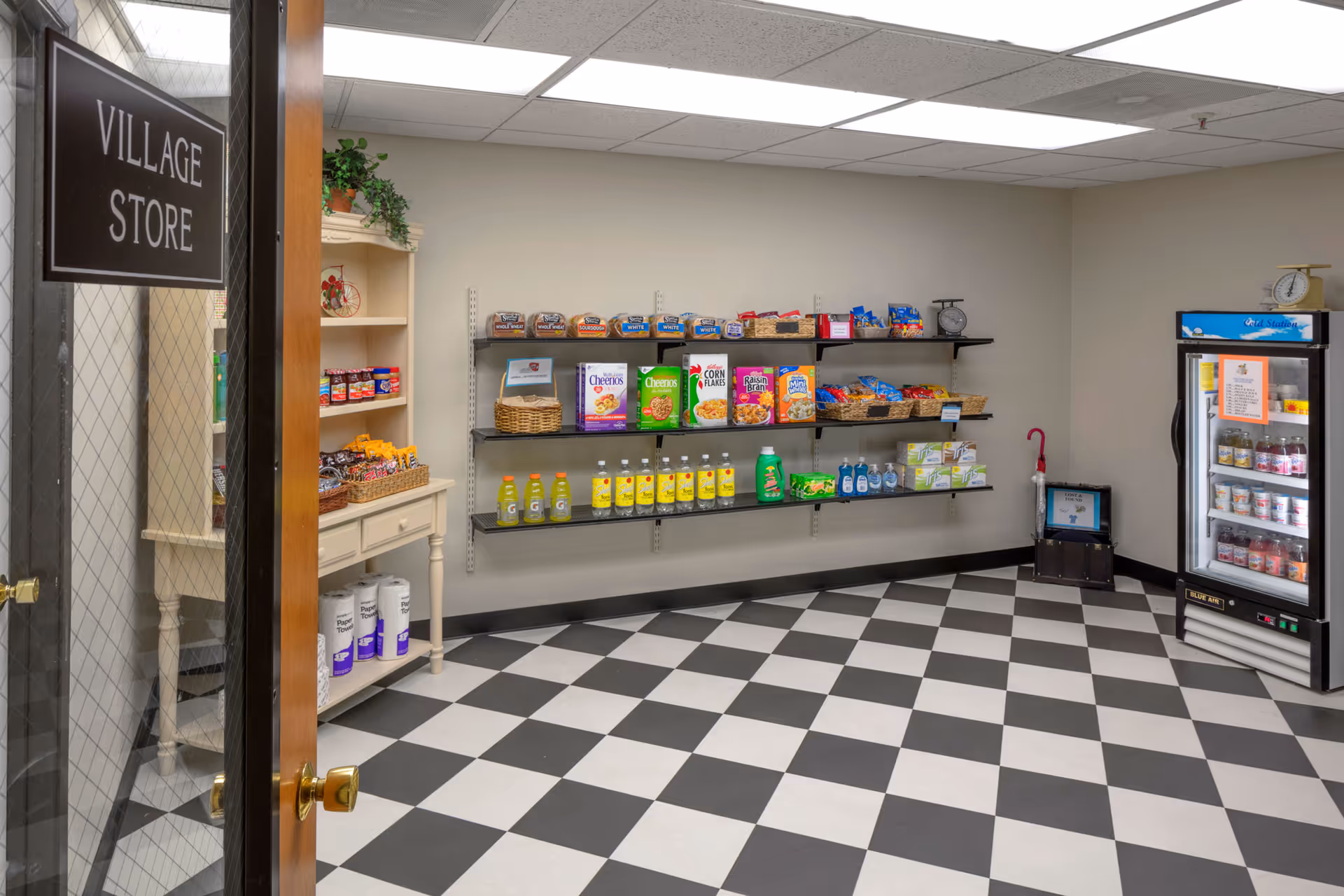 Interior view of a small village store with black and white checkered floor tiles. The store has shelves stocked with various grocery items including cereals, snacks, drinks, and household products. A glass door with a sign reading 'Village Store' is partially open, and a refrigerated display case is visible on the right side of the room.