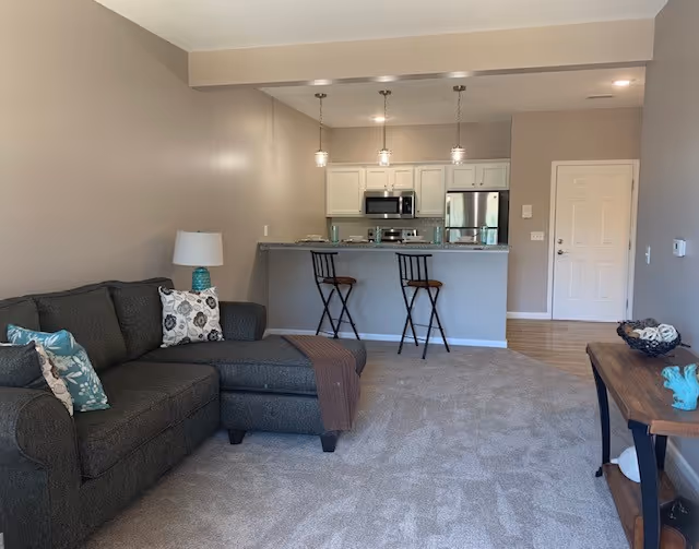 Open-concept living room with a dark gray sectional sofa, carpeted floor, and a kitchen with a breakfast bar and two stools.