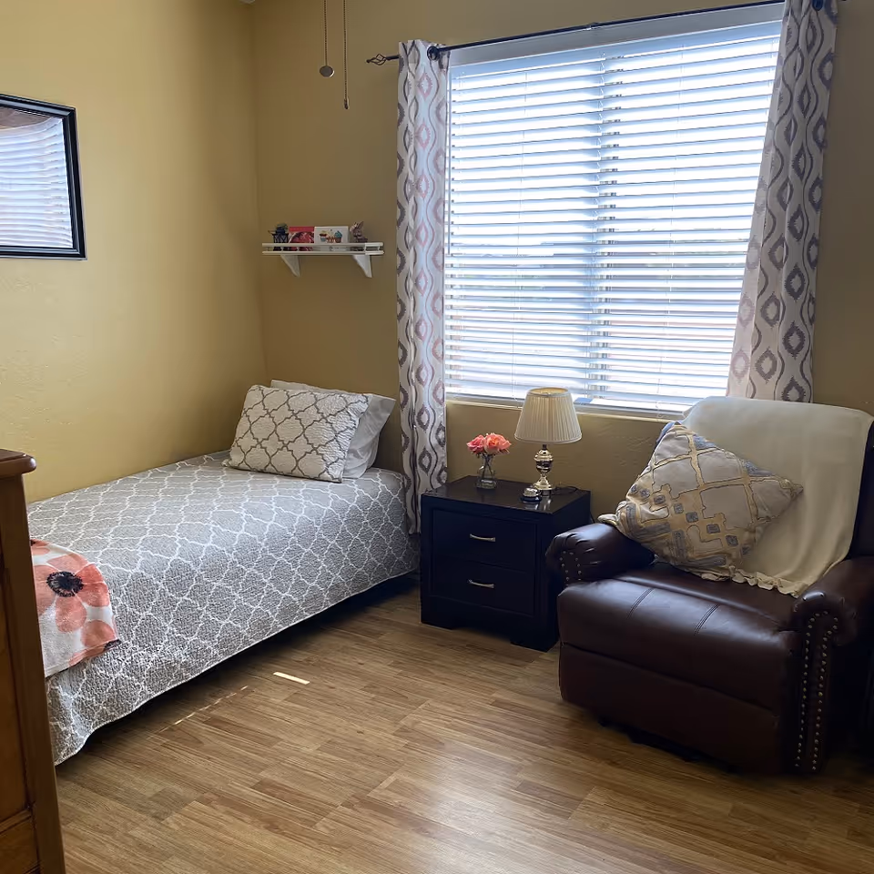 A small, cozy bedroom with a bed, nightstand, chair, and window blinds.