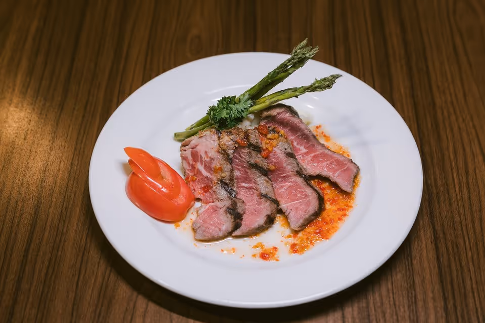 A white plate with slices of medium-rare steak drizzled with sauce, garnished with two asparagus spears, a sprig of parsley, and a tomato carved into a decorative shape, placed on a wooden table.