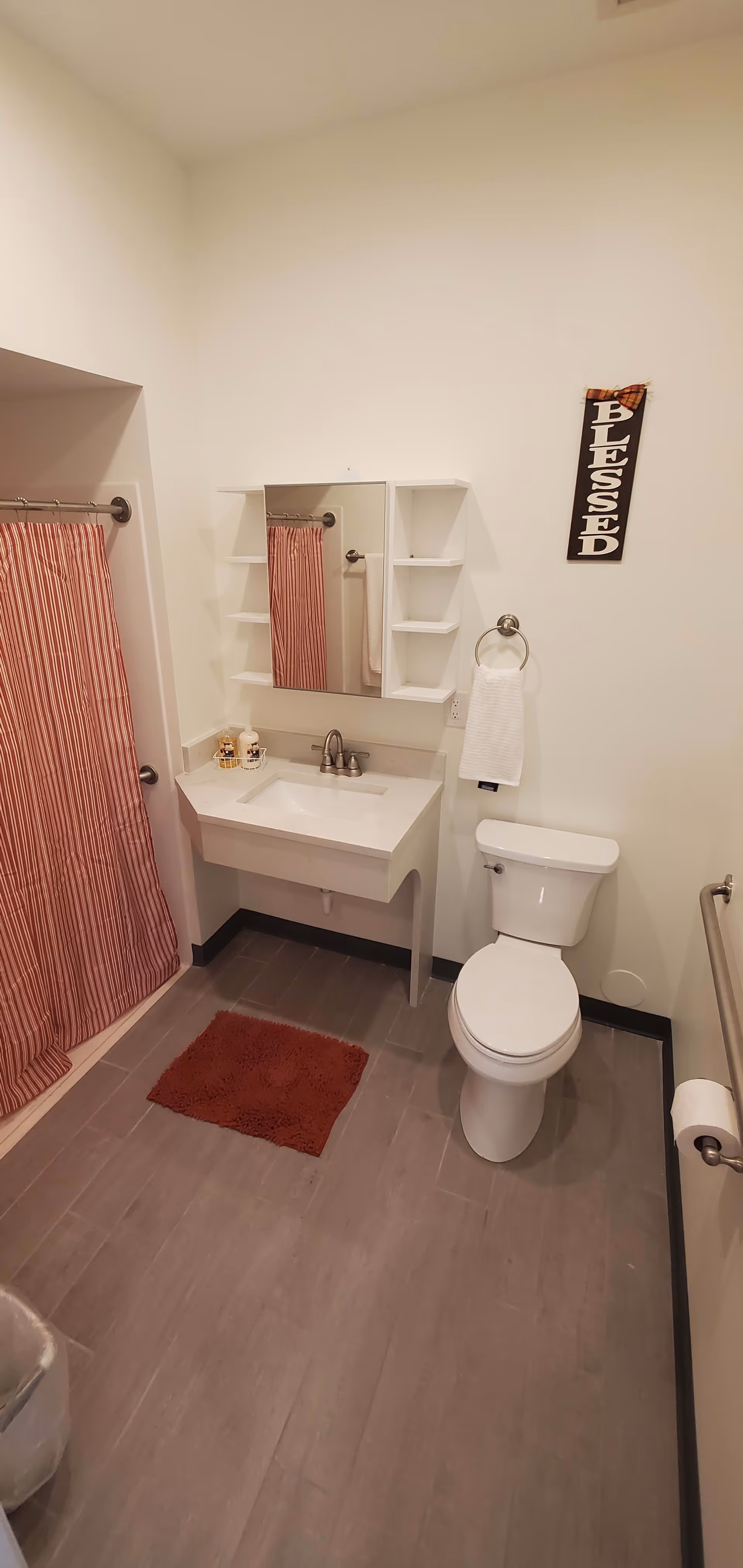 A clean bathroom with a white toilet, a wall-mounted sink with a mirror cabinet above it, a red and white striped shower curtain, a red bath mat on the gray tiled floor, a towel ring with a white hand towel, and a wall decoration with the word 'BLESSED'.