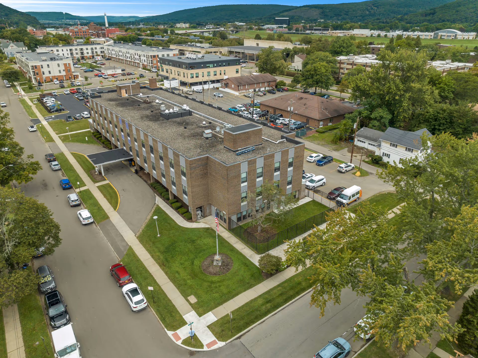 Aerial view of a multi-story brick building with a flat roof, surrounded by parking lots, streets, and greenery. The building is located in a suburban area with other buildings and hills visible in the background. There is a flagpole with an American flag in front of the building.