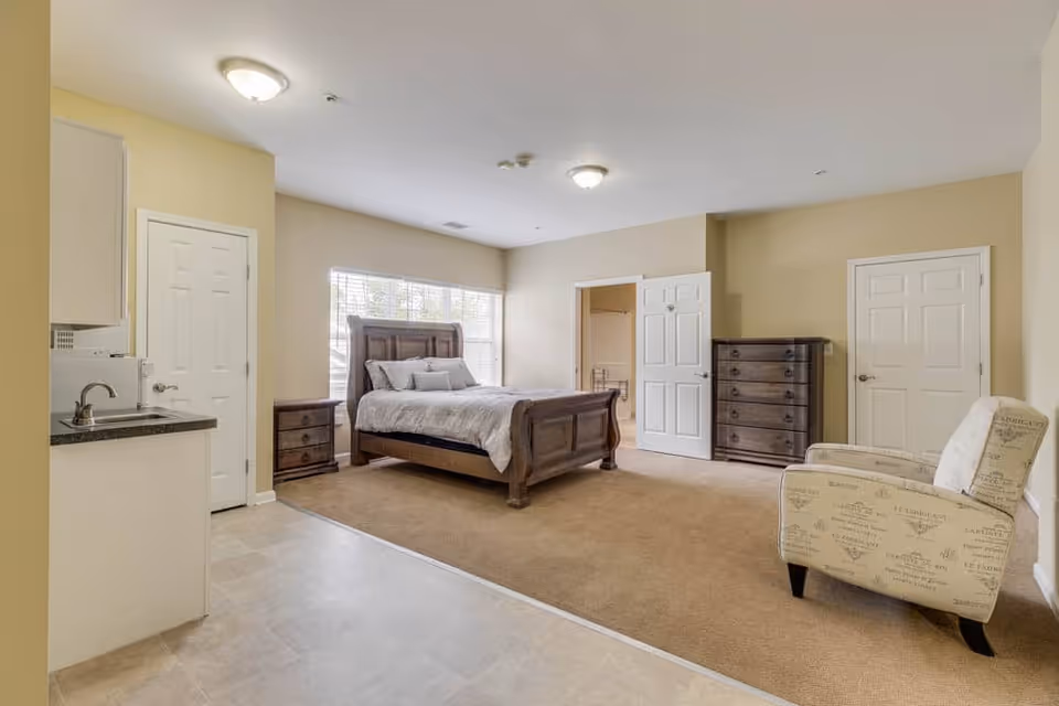 Spacious studio-style bedroom with a wooden bed, dressers, an upholstered armchair, and a small kitchenette on the left.