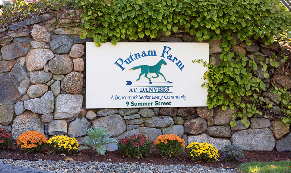 A stone wall with green ivy growing on top and a white sign mounted on it. The sign reads 'Putnam Farm AT DANVERS A Benchmark Senior Living Community 9 Summer Street' with a green horse graphic in the center. Colorful flowers are planted at the base of the wall.