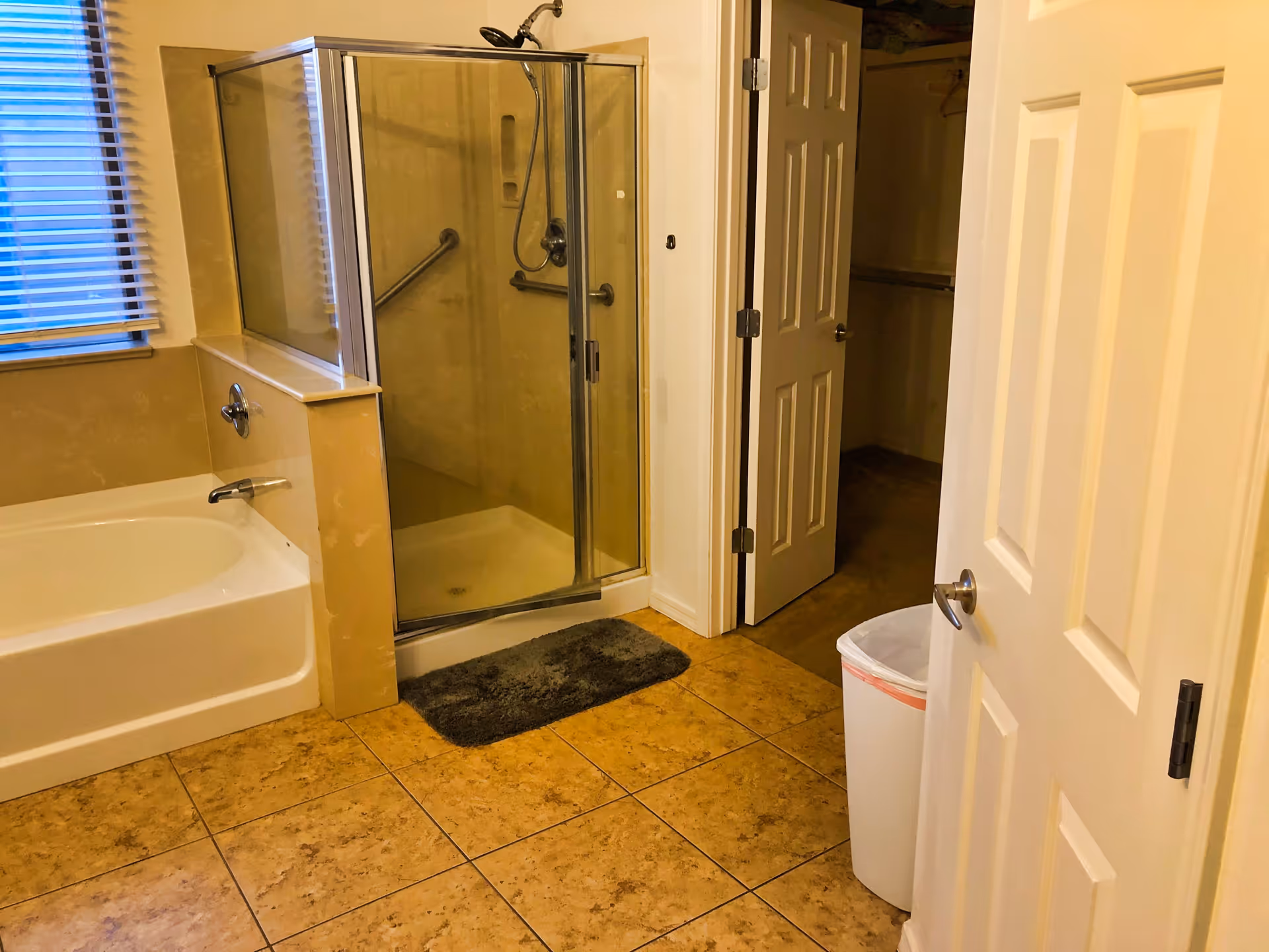 Bathroom with a bathtub, glass-enclosed shower with grab bars, tiled floor, and an open door to a closet.