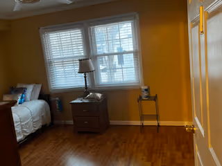 A small bedroom with a single bed on the left side, a nightstand with a lamp next to the bed, a window with blinds letting in natural light, and a small table with a tissue box on the right side. The room has wooden flooring and yellow walls.
