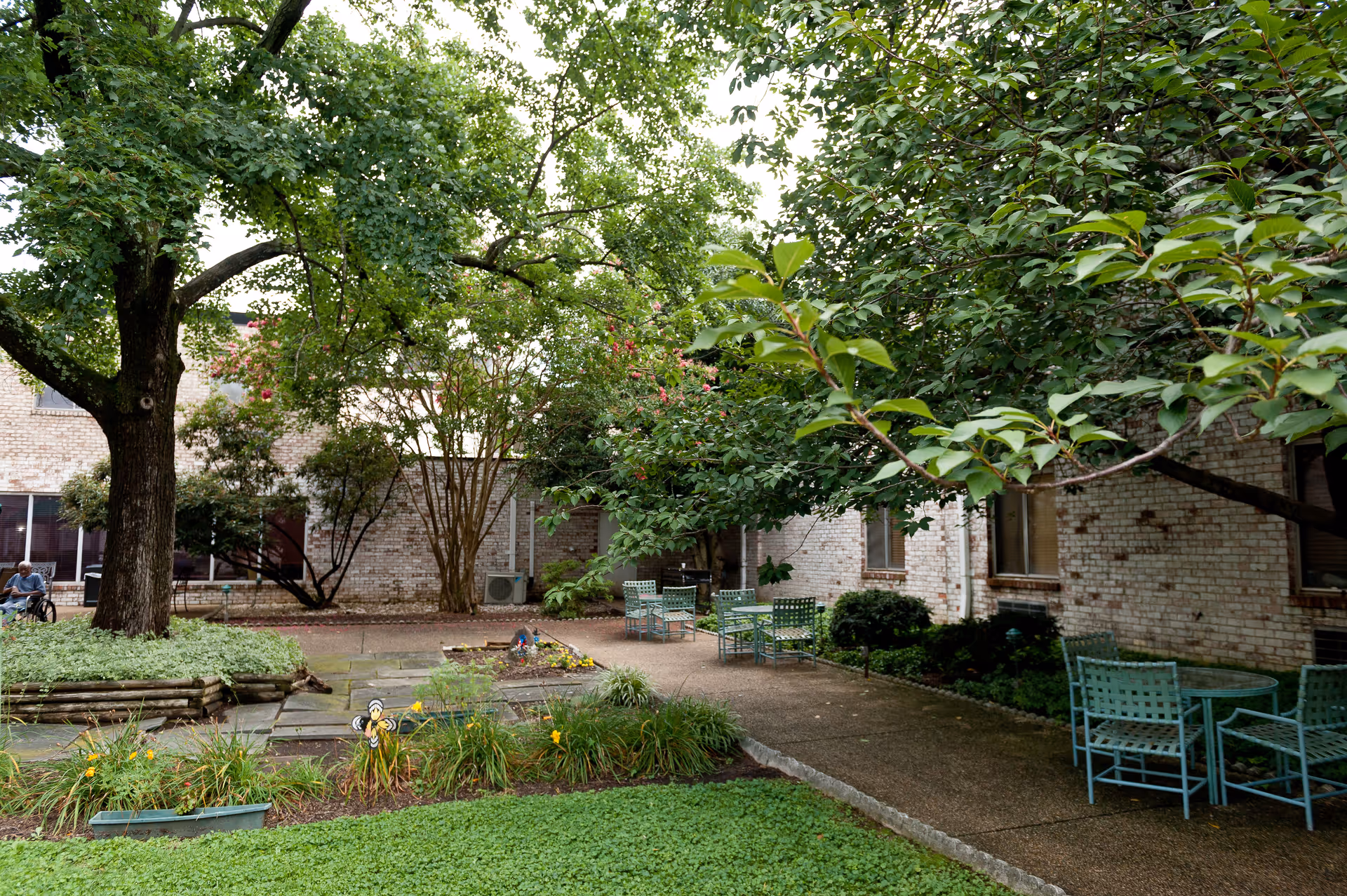Outdoor courtyard area with green trees, shrubs, and flower beds surrounded by brick building walls. Several green metal chairs and tables are placed on the paved walkway. A person in a wheelchair is visible in the background near the building.