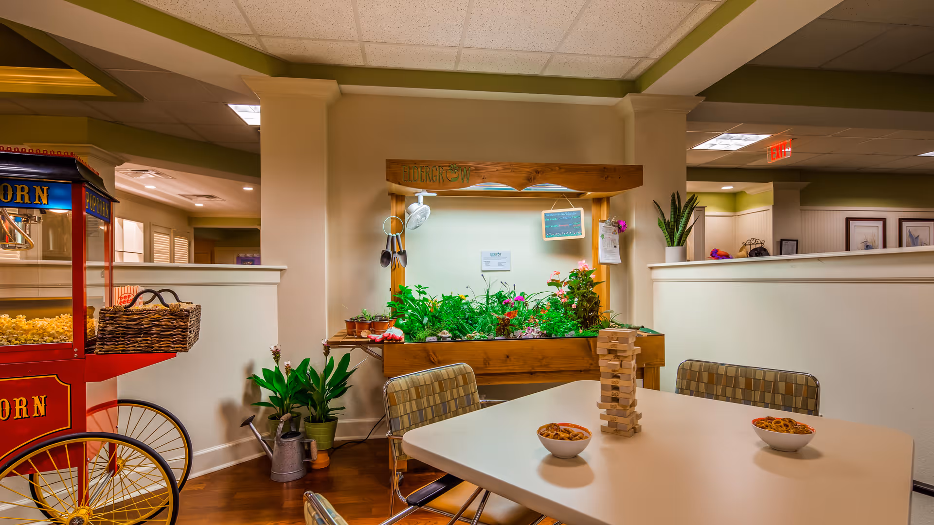 Indoor common area with a white table and four chairs, a Jenga game and two bowls with snacks on the table. Behind the table is a wooden planter box with various green plants and flowers under a grow light. To the left is a red popcorn machine with a basket on top. The room has light-colored walls and a ceiling with recessed lighting.