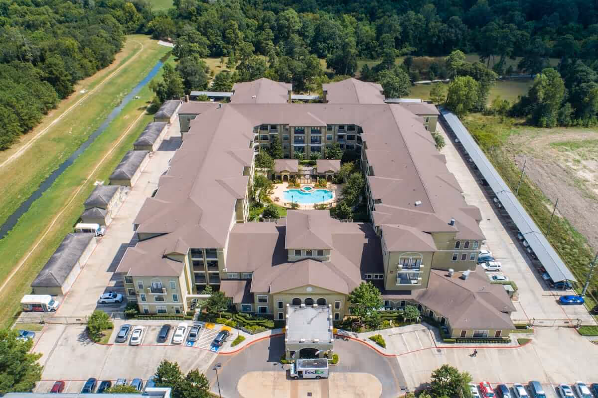 Aerial view of Conservatory At Champion Forest, a large multi-story residential building with a central courtyard featuring a swimming pool and surrounding greenery. The building is surrounded by parking areas and trees, with a FedEx truck visible near the entrance.