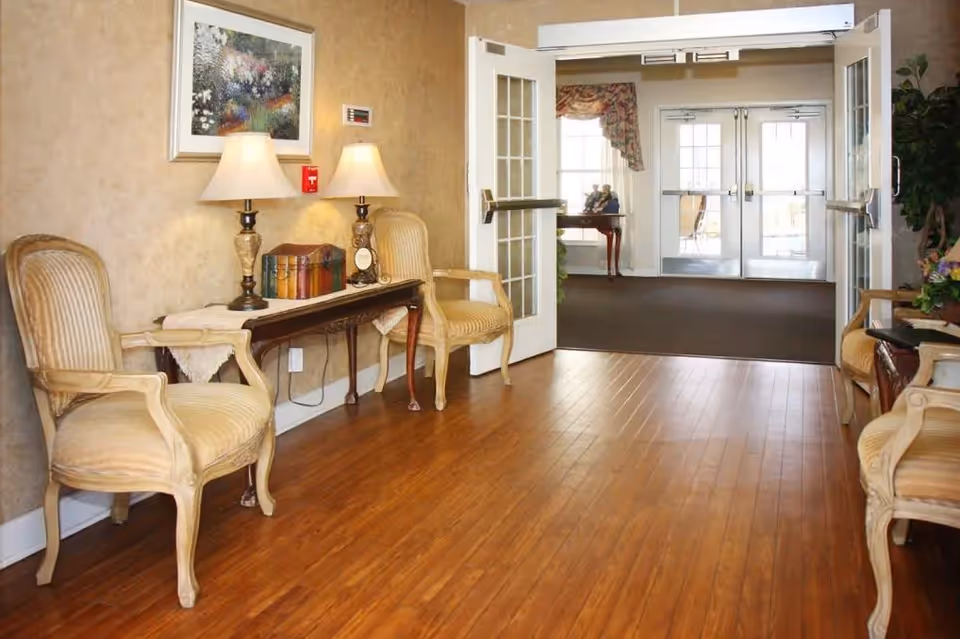 Entrance lobby with wooden floors, paired upholstered armchairs and a console table with lamps leading to double glass doors.
