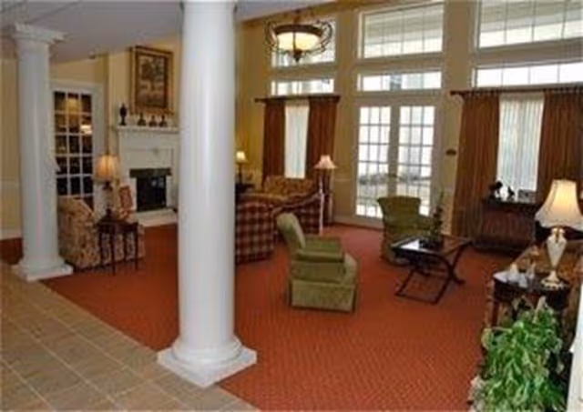 A spacious living room area with large white columns, red carpet, and multiple seating options including green and plaid armchairs. There are several lamps, a fireplace with a framed picture above it, large windows with brown curtains, and a glass door leading outside. The room has a warm and inviting atmosphere.