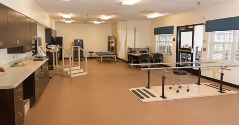A spacious rehabilitation or physical therapy room with parallel bars for walking exercises, therapy tables, chairs, and cabinets along the left wall. The room has beige carpeting, fluorescent ceiling lights, and large windows letting in natural light.