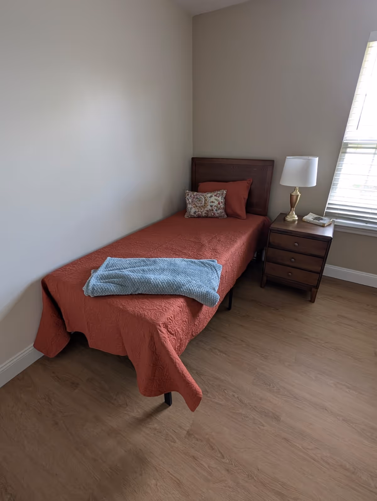 A simple bedroom with a single bed covered in a rust-colored quilt, a blue blanket folded at the foot, and two pillows including one floral decorative pillow. Next to the bed is a wooden nightstand with three drawers, a brass lamp with a white shade, and a book. A window with white blinds is partially visible on the right side, allowing natural light into the room. The floor is wood, and the walls are painted light beige.