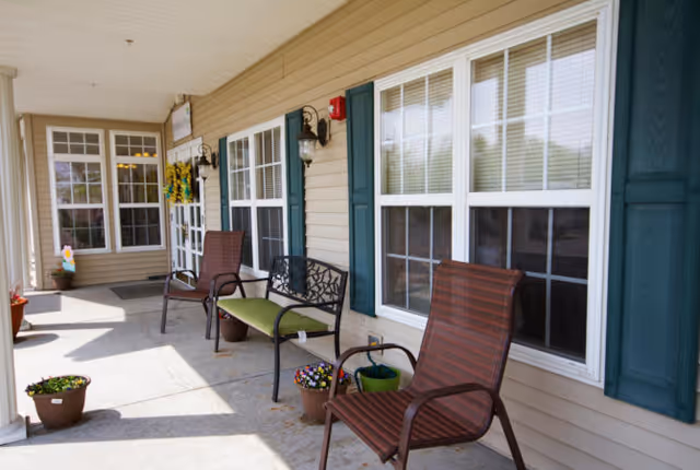 Covered outdoor porch area with beige siding and white-framed windows with blue shutters. The porch has several chairs including a brown striped chair, a black metal bench with a green cushion, and another brown chair. There are potted plants placed along the porch and a door with a wreath hanging on it.