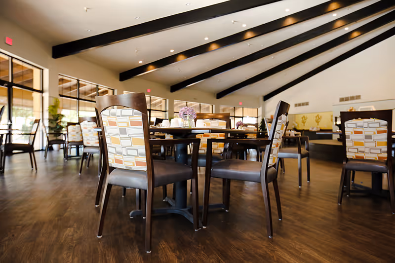 Interior view of a spacious dining room with wooden floors, multiple tables and chairs with patterned upholstery, and large windows letting in natural light. The ceiling has exposed dark beams and recessed lighting.