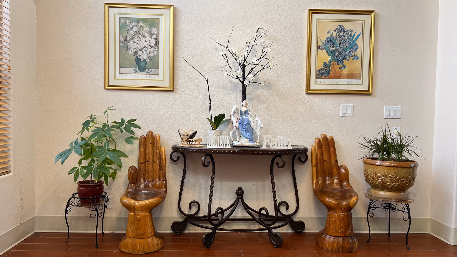 An interior corner of a room featuring a decorative black metal table with a white flowering branch, a figurine, and the words 'BELIEVE' and 'Faith' displayed on top. On either side of the table are two wooden chairs shaped like hands. There are two potted plants on black metal stands, one on the left and one on the right. Above the table hang two framed floral paintings on a beige wall.