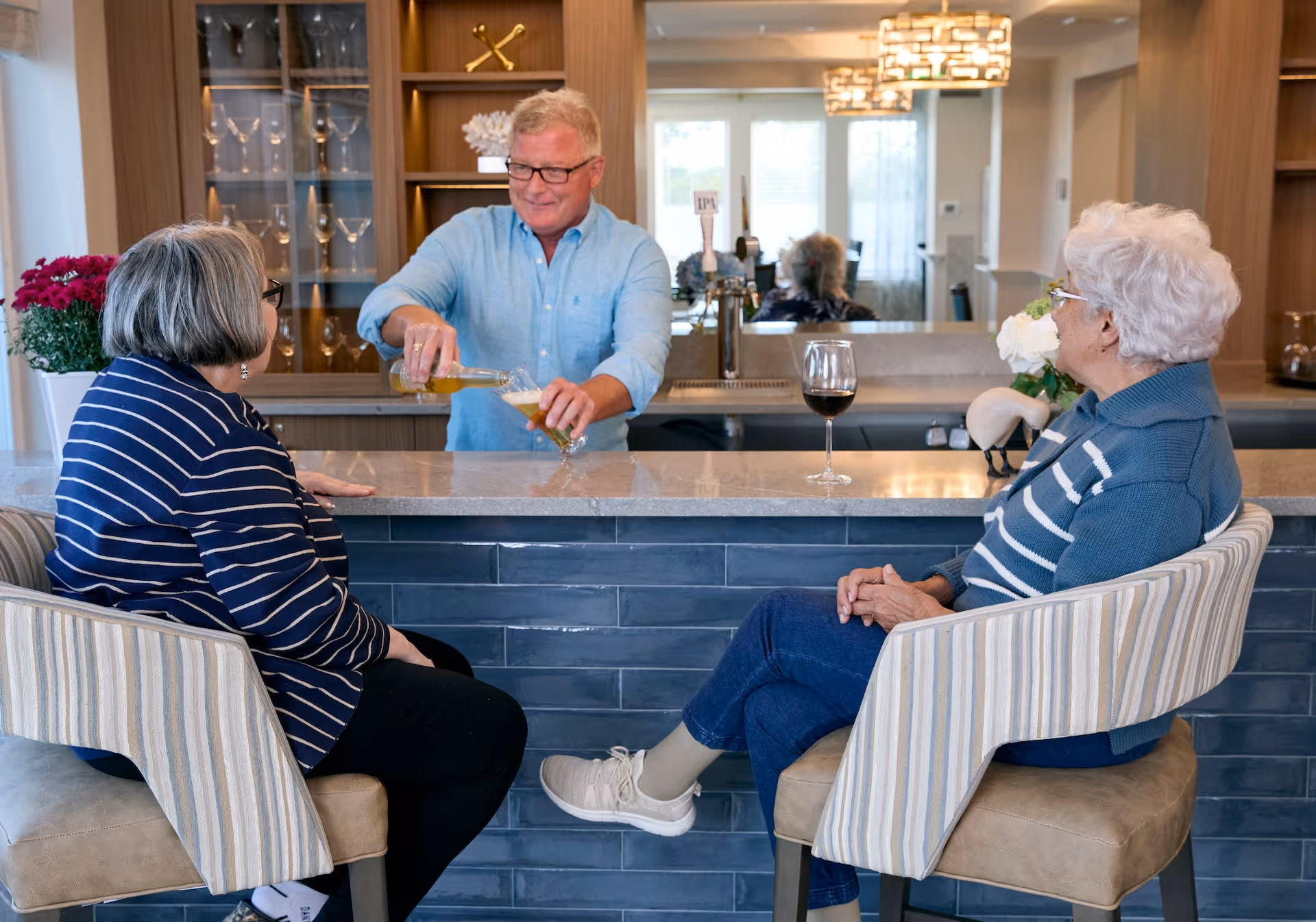 Two elderly women sitting on striped cushioned bar stools at a bar counter, facing a man in a light blue shirt who is pouring a drink into a glass. The background shows a cabinet with glassware and a window letting in natural light.