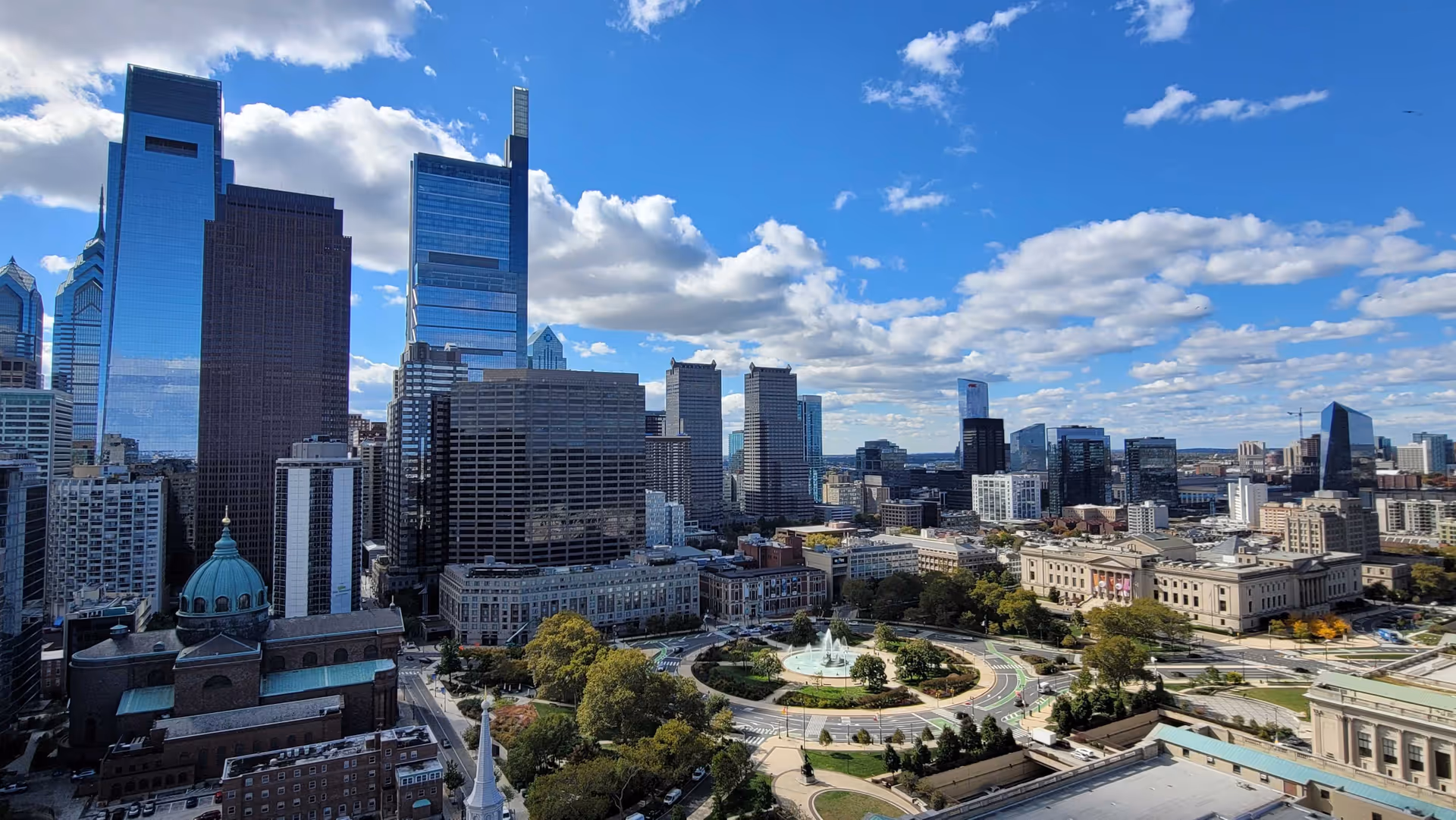 A panoramic view of a city skyline featuring tall modern skyscrapers, a circular park with a fountain in the center, and various other buildings under a partly cloudy blue sky.