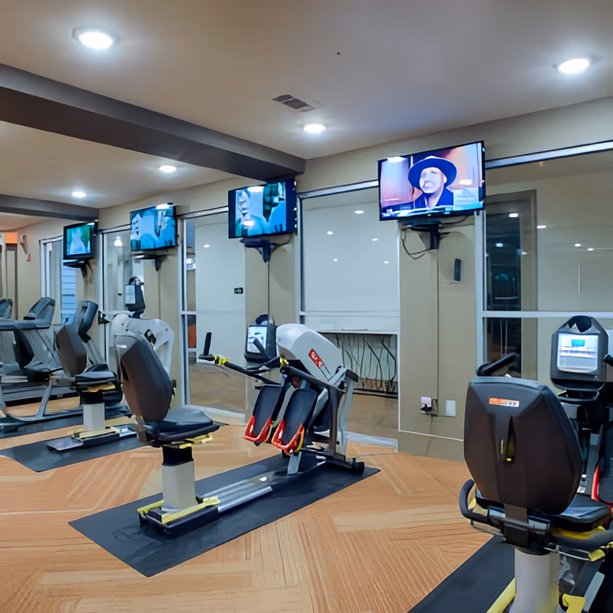 Interior view of a fitness room with exercise machines including recumbent bikes, mounted televisions on the walls, and large mirrors reflecting the equipment and room.