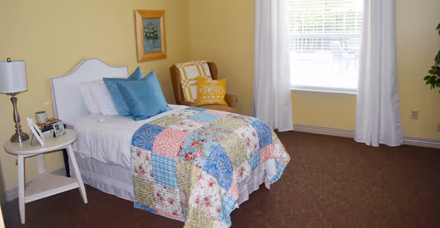 A cozy bedroom with a single bed covered in a colorful patchwork quilt and blue pillows. There is a small round white side table with a lamp, picture frames, and a glass. A wooden chair with a yellow cushion that says 'SWEET HOME' is placed near a window with white curtains. The walls are painted light yellow and the floor is carpeted in brown.