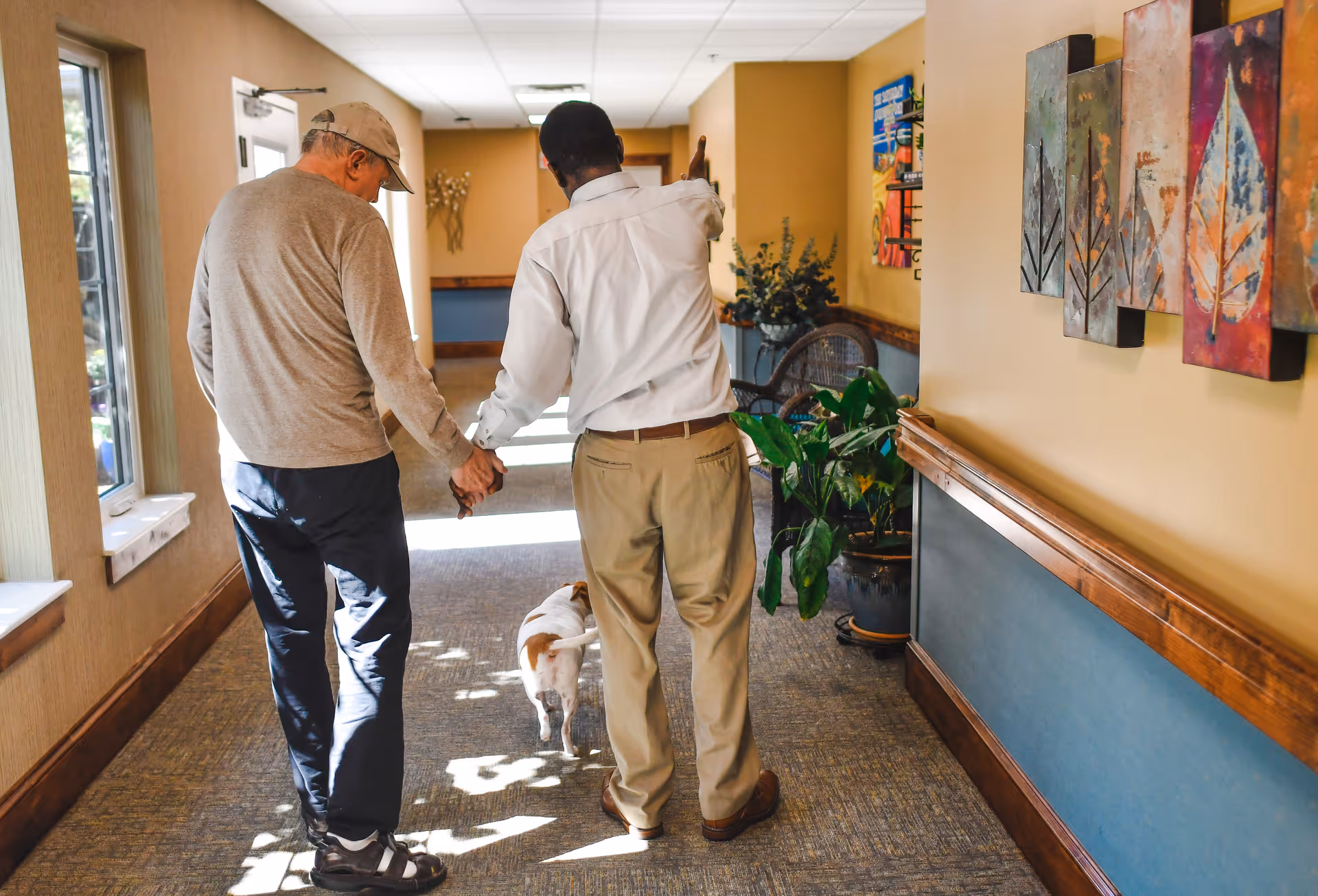 Two men walking down a hallway inside a facility, holding hands. One man is elderly wearing a cap, gray sweater, and dark pants, while the other man is younger, wearing a white shirt and khaki pants. A small dog walks ahead of them. The hallway has windows on the left, plants, and colorful artwork on the right wall.